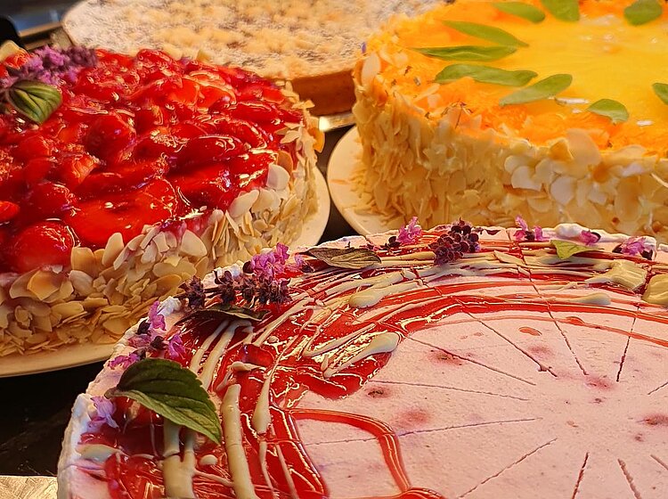 Drei dekorierte Kuchen auf einem Tisch, einer mit roter Fruchtsauce und Blüten, einer mit Mandeln und einer mit gelber Glasur und grünen Blättern. Im Hintergrund ist unscharf ein Café mit Tischen und Stühlen zu sehen.