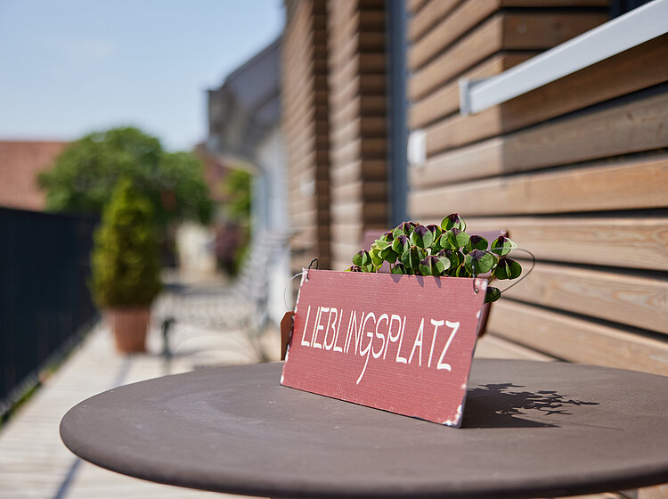 Ein Schild mit der Aufschrift Lieblingsplatz steht auf einem runden Tisch vor einer Holzwand, im Hintergrund unscharfe Pflanzen und Sitzbänke auf einem Balkon.