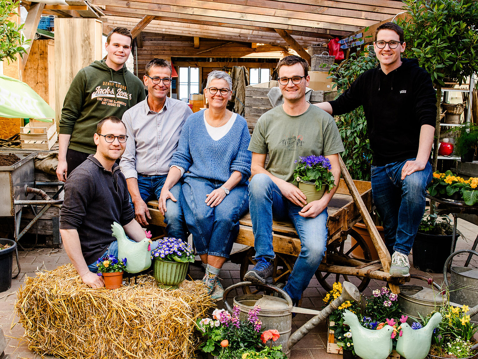 Sechs Menschen, vier Männer und zwei Frauen, sitzen und stehen in einem Gewächshaus um eine Heuballen und bunte Blumentöpfe. Sie lächeln und wirken entspannt in einer Gartenumgebung mit Pflanzen und Gartengeräten.